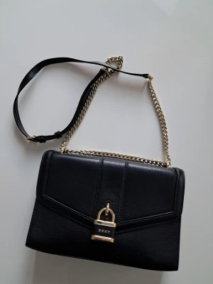 Svart axelväska från DKNY med kedja - Snygg svart axelväska från DKNY i slätt läder med guldfärgad kedjerem och coolt lås framtill. Väskan har en stilren, rektangulär form och detaljer i guld som ger en lyxig känsla. Perfekt för dig som vill ha en trendig och tidlös accessoar. Obs! Som ny. 