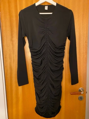 Svart långärmad omlottklänning - Snygg svart klänning med långa ärmar och rynkade detaljer längs hela framsidan och baksidan. Klänningen har en kroppsnära passform och v-ringning. Perfekt för dig som vill ha en stilren men ändå unik look.