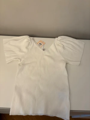 Vit topp - Vit ribbad topp från Only med v-ringning och korta puffärmar. Toppen har en smickrande passform och är tillverkad i ett mjukt, stretchigt material som känns bekvämt mot huden. Perfekt för att lyfta vilken outfit som helst.