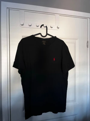 Svart t-shirt från Polo Ralph Lauren - Klassisk svart t-shirt från Polo Ralph Lauren med röd broderad logga på bröstet. Tillverkad i 100% bomull och har en normal passform med korta ärmar. Perfekt basplagg för en stilren look.