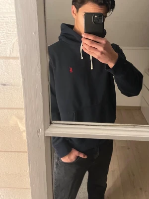 Mörkblå hoodie från Polo Ralph Lauren - Mörkblå hoodie från Polo Ralph Lauren med klassisk röd logga broderad på bröstet. Hoodien har vit dragsko, känguruficka och mjukt material som känns skönt mot huden. Perfekt för en avslappnad och stilren look.