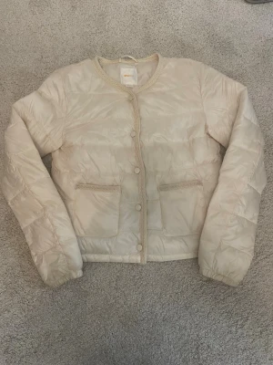 Beige/vit Gina tricot pufferjacka - Säljer en beige pufferjacka med rund halsringning och tryckknappar framtill. Jackan har två stora framfickor och quiltad design. Perfekt för våren och enkel att matcha med olika outfits ❤️
