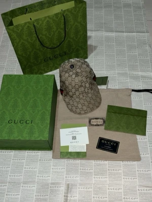 Gucci GG Supreme keps beige - Snygg keps från Gucci i klassiskt beige GG Supreme-mönster med bruna detaljer och röd-grön rand på sidan. Kepsen har böjd skärm och justerbar rem bak. Tillverkad i canvasmaterial med läderdetaljer. Perfekt för dig som vill sticka ut med lyxig streetstyle.