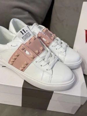Valentino Rockstud Untitled sneakers - Snygga vita sneakers från Valentino med ljusrosa band och ikoniska nitar över vristen. Skorna har en clean design med vita snören och låg profil. Perfekta för dig som vill ha en lyxig och edgy touch till din outfit. Materialet är slätt läder och detaljerna ger en exklusiv känsla.
