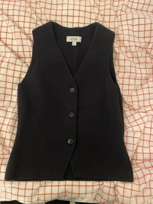 Svart väst med knappar från COS - Stilren svart väst från COS med tre knappar framtill och djup V-ringning. Västen har en rak passform och är ärmlös, perfekt för att lägga till en clean look till din outfit. Materialet ser ut att vara mjukt och något tjockare, vilket ger en exklusiv känsla.