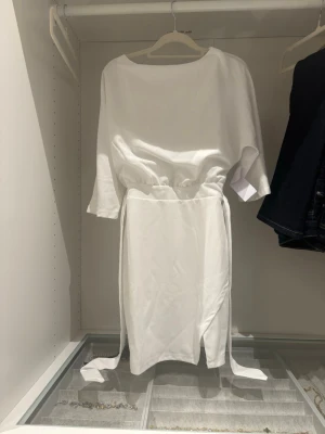 Vit midiklänning med knytband - Stilren vit midiklänning med trekvartsärmar och rund halsringning. Klänningen har resår i midjan och långa knytband i sidorna för en justerbar passform. Tillverkad i ett mjukt, lätt tyg som ger en avslappnad men ändå chic look.