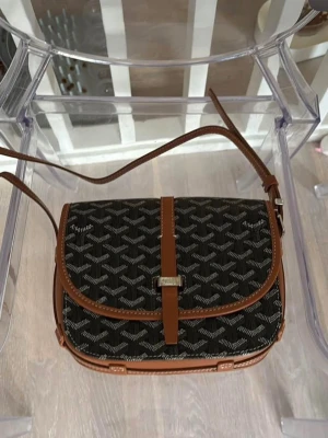  svart axelväska med mönster - Snygg svart axelväska från  med klassiskt vitt mönster och bruna läderdetaljer. Väskan har justerbar axelrem och silverfärgat spänne framtill. Perfekt storlek för det viktigaste och en tidlös design som sticker ut.
