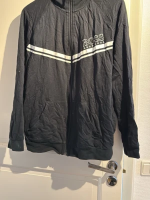 Svart ziptröja från Hugo Boss - Snygg svart ziptröja från Hugo Boss med vita ränder över bröstet och logga på vänster sida. Tröjan har hög krage, dragkedja framtill och fickor på sidorna. Perfekt för en sportig och avslappnad stil.