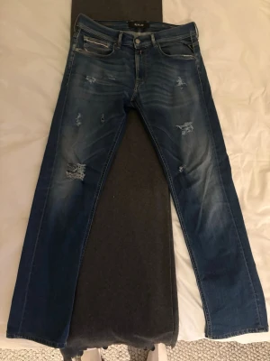 Blå slitna jeans från Replay - Snygga blå jeans från Replay med slitningar och distressed detaljer på både fram- och baksida. Klassisk femficksmodell med raka ben och tydliga tvättade partier. Perfekta för en avslappnad och trendig look.
