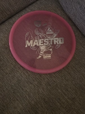 Discmania Maestro discgolf disc - Säljer en rosa Discmania Maestro discgolf disc i plast, mid-range modell med tryckt motiv och text. Märkt med Active Premium och flight numbers 4/3/0/2. Perfekt för dig som vill ha kontroll på banan.