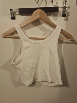 Vit croppad linnetopp - Säljer en enkel och stilren vit croppad linnetopp i ribbad bomull. Toppen är ärmlös med breda axelband och rundad halsringning, perfekt för varma dagar eller som lager under skjortor.