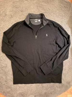 Svart halfzip från Polo Ralph Lauren - Svart långärmad tröja från Polo Ralph Lauren med halv dragkedja och klassisk broderad logga på bröstet. Tröjan har ståkrage med grå insida och är tillverkad i mjukt bomullsmaterial. Inte använd mycket så fint skick!