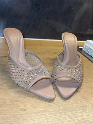  Beiga Klackar som passar till dejter - Säljer ett par snygga slip-in sandaler i beige med nätmönster och glittriga stenar på ovandelen. Sandalerna har öppen tå och låg klack, samt en modern fyrkantig tåform. Perfekta för dig som vill ha något trendigt och bekvämt.