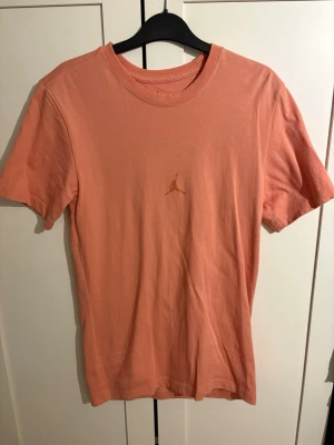 Jordan t-shirt i korall med tryck - Snygg korallfärgad t-shirt från Jordan med klassisk Jumpman-logga på bröstet och stort 23-tryck med signatur på ryggen. Tillverkad i mjuk bomull och har en normal passform med korta ärmar.