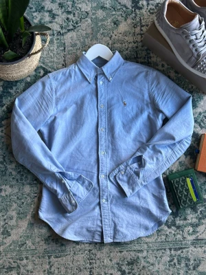 Ljusblå skjorta från Ralph Lauren - Klassisk ljusblå skjorta från Ralph Lauren med button-down krage och broderad logga på bröstet. Skjortan har super slim fit passform, långa ärmar och knappar framtill. Perfekt för en clean och stilren look.