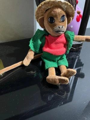 Mjuk apa med halmhatt och gröna kläder - En söt mjukisdocka i form av en apa med halmhatt, grön skjorta och shorts samt röd tröja. Apan har långa armar, ben och svans, och är tillverkad i mjukt tyg. Perfekt som dekoration eller leksak.