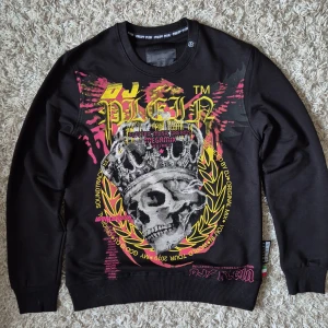 Svart Philipp Plein sweatshirt S i nyskick  - Svart sweatshirt med ett färgstarkt grafiskt tryck fram och bak, kristallglittrande döskalle med krona och text i gult, rosa och vitt. Rund hals, ribbade muddar och lång ärm. Materialet är mjuk bomull, perfekt för dig som gillar statement-plagg. 
