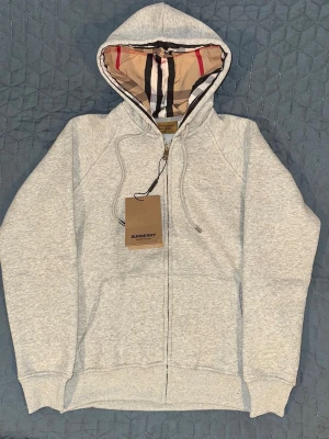 Burberry Grå zip-hoodie  - Snygg grå hoodie från Burberry med dragkedja och klassiskt rutigt foder i huvan. Tröjan har dragsko i huvan, ribbade muddar och två fickor framtill. Perfekt för dig som gillar exklusiva streetwear-plagg med stilren design.