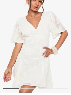 Vit studentklänning från Boohoo - Söt vit kortklänning från Boohoo med omlott och v-ringning. Klänningen har korta, vida ärmar och subtilt blommigt mönster i tyget. Superfin och oanvänd! Storlek M