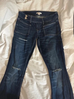 Mörkblå slitna jeans från Gina Tricot - Snygga mörkblå jeans från Gina Tricot med slitningar på knäna och framsidan. Jeansen har utsvängda ben, dubbla knappar i midjan och stora fickor fram. Perfekta för en cool och avslappnad stil. De är i tall (ca 84 i innerbenslängd)