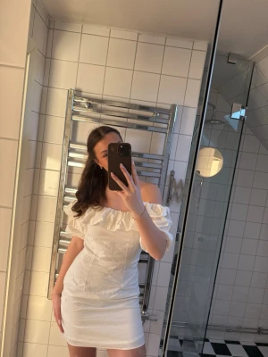 Vit offshoulder kortklänning med volang - Snygg vit kortklänning med offshoulder och volangdetalj upptill. Klänningen har en figurnära passform och är perfekt för sommarens tillfällen. 