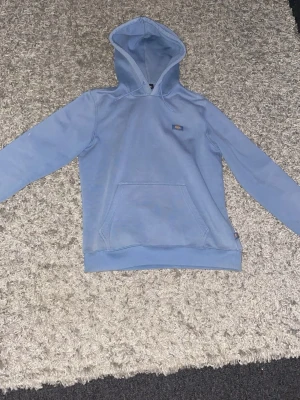 Ljusblå hoodie från Dickies - Snygg ljusblå hoodie från Dickies med klassisk känguruficka och liten logotyp på bröstet. Mjuk bomullskänsla och ribbade muddar vid ärmslut och nederkant. Perfekt för en avslappnad streetwear-look.