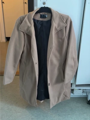 Beige kappa med svarta knappar - Stilren beige kappa med svart foder och knäppning framtill. Kappan har bred krage och fickor på sidorna. Ger en tidlös look till din outfit. Köpt för 399kr, Storlek XL, Samt aldrig använd. Beige mer mot de bruna hållet.