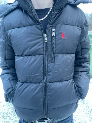 Svart pufferjacka från Ralph Lauren - Säljer en svart pufferjacka från Ralph Lauren med röd broderad logga på bröstet. Jackan har huva, dubbla dragkedjor framtill och en bröstficka med dragkedja. Fylld med vaddering för extra värme och har en modern, boxig passform.