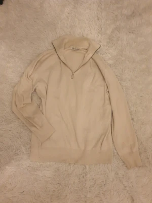 Beige halvzip-tröja från Zara - Snygg beige tröja från Zara med halv dragkedja vid kragen. Tröjan har långa ärmar, ribbade muddar och är tillverkad i ett mjukt stickat material. Perfekt för lager på lager och enkel att matcha med olika outfits.
