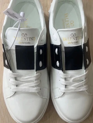Valentino Garavani vita sneakers - Snygga vita sneakers från Valentino Garavani med svart bred rem och dekorativa nitar på sidorna. Skorna har klassisk låg profil, vita skosnören och guldfärgad logotyp på plösen. Tillverkade i slätt läder för en clean och lyxig look.