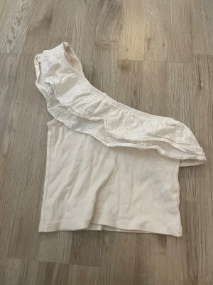 Vit oneshoulder topp från Gina Tricot - Snygg vit oneshoulder topp från Gina Tricot med bred volang i broderad spets. Toppen är ribbad och har en axel bar, vilket ger en trendig look. Perfekt för dig som vill sticka ut lite extra.