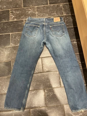 Levi's 501 - Klassiska blå jeans från Levi's med rak passform och knappgylf. Jeansen har fem fickor och tydliga sömmar, samt den ikoniska läderlappen bak i midjan. Perfekta för en avslappnad och tidlös stil.