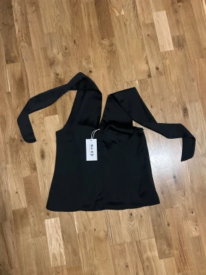 Lyxig satintopp från NA-KD - Trendig satin topp från NA-KD i slätt, glansigt material. Toppen har breda band som knyts över axlarna och en djup v-ringning framtill. Oanvänd med prislapp kvar på. 