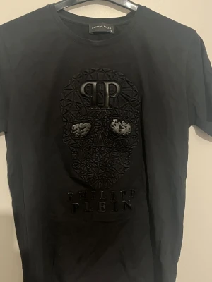 Svart t-shirt med döskalle från Philipp Plein - Cool svart t-shirt från Philipp Plein med 3D-döskalle och logotyp framtill. T-shirten har rund hals och är tillverkad i mjukt material. Perfekt för dig som gillar streetstyle och detaljer som sticker ut. Storlek s men passar xs 