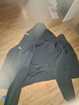 Svart vindjacka från Quiksilver - Snygg svart vindjacka från Quiksilver med klassisk logga på ärmen. Jackan har huva och dragkedja framtill. Tillverkad i ett lätt och slitstarkt material som passar perfekt för blåsiga dagar. Enkel och stilren design som funkar till det mesta.