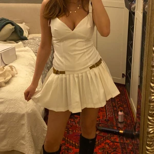 Vit kort plisserad klänning med bälte - Supersöt vit kort klänning med plisserad kjol och smala axelband. Klänningen har en v-ringad urringning och två bruna bälten vid midjan som ger en skitsnygg detalj. Klänningen finns inte kvar på hemsidan, köpt för 500kr 💕