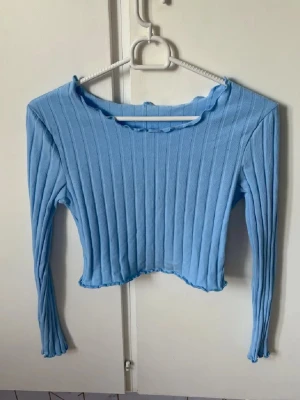 Ljusblå ribbad croppad topp - Trendig ljusblå croppad topp med lång ärm och ribbad struktur. Toppen har en lätt vågig kant vid halsringning och nederkant för en extra detalj. Perfekt för att styla med höga jeans eller kjol.