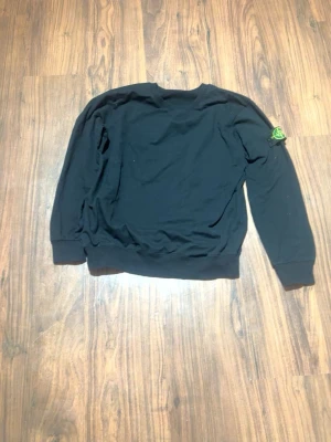 Svart sweatshirt från Stone Island - Svart sweatshirt från Stone Island med klassisk rund hals och ribbade muddar. Ikonisk logotyp-patch på vänster ärm. Perfekt för en stilren och avslappnad look.