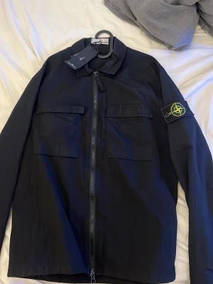 Svart overshirt från Stone Island - Svart overshirt från Stone Island med klassisk kompasspatch på vänster ärm. Jackan har två stora bröstfickor med lock och dragkedja framtill. Tillverkad i ett lätt, slitstarkt material som ger en clean och modern look.