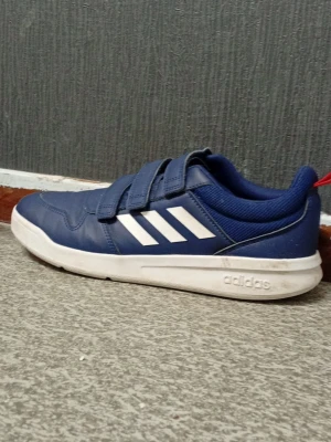 Blå Adidas sneakers med kardborre - Snygga blå sneakers från Adidas med vita sulor och tre kardborreband för enkel påtagning. Skorna har en röd detalj på plösen och klassisk Adidas-logga under sulan. Ovandelen är i syntetmaterial och textil för en sportig look. Skorna har bara använts en gång för o testa inomhus den var för stor och aldrig använts mer STORLEK 40