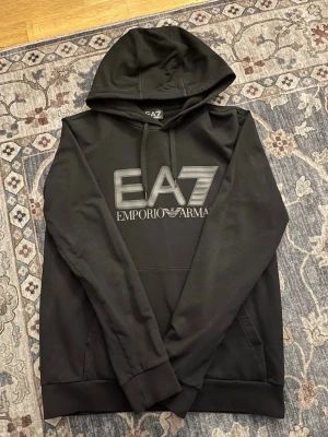 Svart hoodie från EA7 Emporio Armani - Snygg svart hoodie från EA7 Emporio Armani med stor logga framtill. Klassisk modell med huva och dragsnören samt stor ficka på magen. Tillverkad i mjukt material som känns skönt mot huden. Perfekt för en avslappnad och trendig look.
