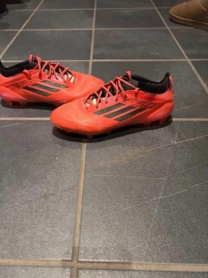 Adidas F50 elite - Snygga röda adidas F50 fotbollsskor med svarta detaljer och klassiska tre ränder på sidan. Skorna har snörning och är tillverkade i syntetmaterial för lätt känsla på planen. Perfekta för dig som vill ha snabbhet och stil på fotbollsplanen.