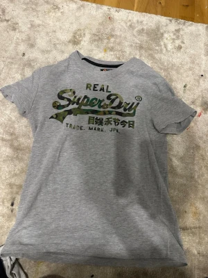 Grå Superdry t-shirt med camologga - Snygg grå t-shirt från Superdry med korta ärmar och ett stort kamouflagemönstrat Superdry-tryck på bröstet. Tillverkad i mjuk bomull, perfekt för en avslappnad och trendig stil.