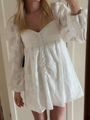 Vit kort klänning med puffärmar - Superfin vit kort klänning med v-ringning, puffiga långärmar och knappar framtill från Gina Tricot. Klänningen har ett subtilt blommigt mönster i tyget och en romantisk känsla. Perfekt för student eller sommarens tillfällen!