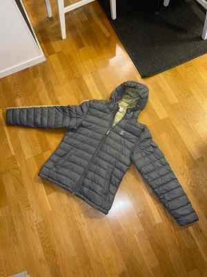 Grå pufferjacka från Dolomite - Säljer en grå pufferjacka från Dolomite med huva och dragkedja. Jackan har quiltad design, två sidofickor och snygga patchdetaljer med italienska flaggan. Insidan är ljusgul och materialet är syntet. Perfekt för kalla dagar!