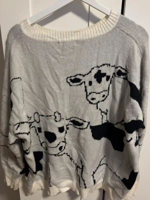 Grå stickad tröja med ko-tryck - Unik grå stickad tröja med svart och vit ko-motiv över hela framsidan. Tröjan har bred ribbad halsringning och muddar vid ärmslut och nederkant. Storlek står inte men skulle säga L