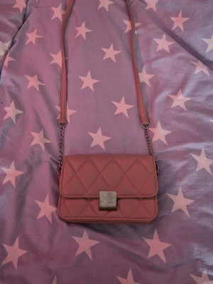 Rosa quiltad axelväska med kedja - Säljer en snygg rosa axelväska med quiltat mönster och silverfärgad kedjedetalj på axelremmen. Väskan har ett stilrent metallspänne framtill och ett rymligt fack med innerficka som stängs med dragkedja. Perfekt storlek för det viktigaste och ger en trendig touch till din outfit.