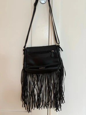 Svart axelväska med fransar från s.Oliver - Säljer en svart axelväska från s.Oliver i syntetmaterial. Väskan har långa fransar längs nederkanten, justerbar axelrem och en dragkedjeficka framtill. .