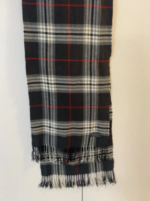 Rutig ullscarf från Burberrys - Säljer en klassisk rutig scarf från Burberrys i svart, grått, vitt och röda detaljer. Scarfen är tillverkad i 100% lammull och har fransar längst ner. Perfekt för dig som gillar att hålla sig varm under vintern.
