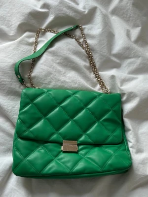Grön quiltad axelväska med kedja - Säljer en snygg grön axelväska med quiltat mönster och guldfärgad kedjerem. Väskan har ett stilrent metallspänne framtill och är tillverkad i skinnimitation. Perfekt för dig som vill ha en färgklick till din outfit!